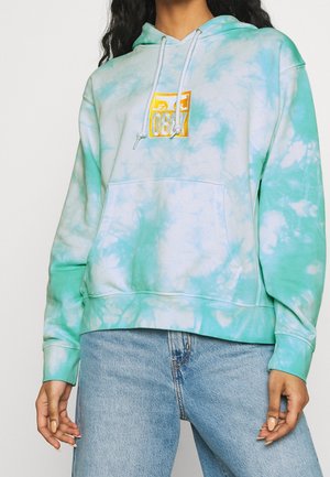 Personne portant un sweat à capuche tie-dye bleu sarcelle et blanc avec un logo OBEY jaune sur la poitrine, et un jean en denim bleu clair, debout avec les mains détendues.