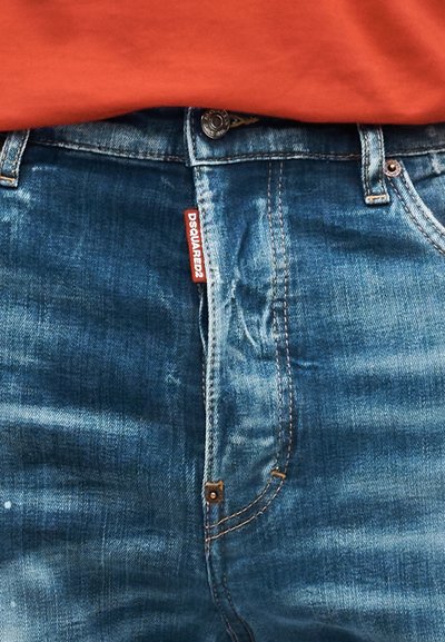 Κοντινή λήψη μπλε τζιν παντελονιού με πορτοκαλοκόκκινη ετικέτα λογότυπου DSQUARED2 και πορτοκαλί πουκάμισο που είναι βιδωμένο στη μέση.