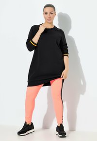 Sweatshirt nero oversize con polsini a strisce arancioni, abbinato a leggings fucsia neon aderenti con strisce laterali nere e sneaker nere.