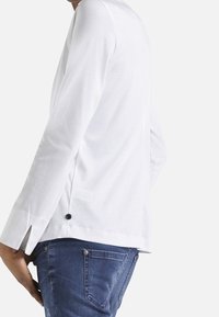 Camicia bianca a maniche lunghe con spacchi laterali e dettagli in bottone neri, abbinata a jeans blu, evidenziando linee semplici e una trama di tessuto liscia.