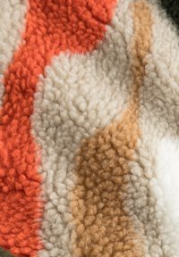 Textúrovaná tkanina s vlnitým vzorom v oranžových, smotanových a svetlohnedých odtieňoch, so nadýchaným a mäkkým povrchom.