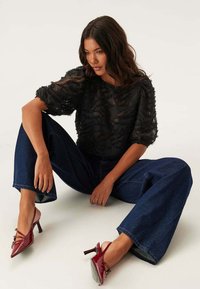 Blusa nera trasparente con motivo floreale testurizzato e maniche a sbuffo, abbinata a jeans in denim a gamba larga e scarpe rosse con tacco e cinturino alla caviglia.
