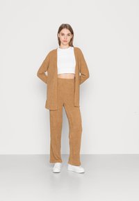 Cardigã de malha castanho claro sobre um topo cropped branco, combinado com calças de malha a condizer. Ténis brancos completam o look.