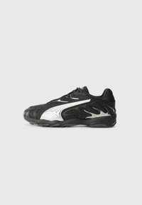 Zapatilla deportiva negra con acentos plateados, parte superior texturizada y suela acolchada. Presenta elementos de diseño curvados y un sistema de cordones.
