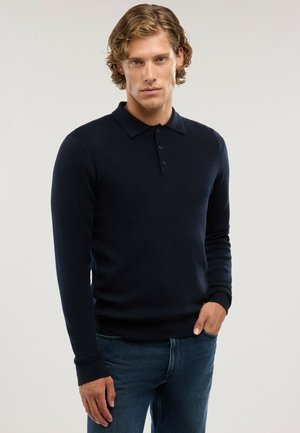 Joven con cabello rubio ondulado que lleva una camisa polo de manga larga azul marino oscuro y vaqueros azules, de pie con una mano en el bolsillo.