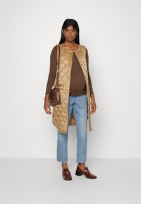 Colete bege acolchoado com decote arredondado e fecho de botões, sobre uma blusa castanha. Combinado com jeans azul claro e mocassins castanhos.