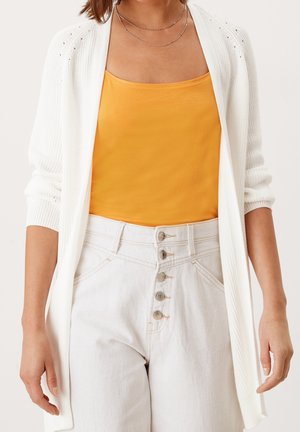 Kvinde iført hvide højtaljede jeans med knapper, en gul top og en hvid åbenstrikket cardigan med opsmøgede ærmer.