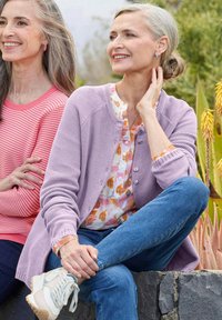 Lichtpaarse gebreide cardigan over een bloemenblouse, gecombineerd met blauwe jeans en witte sneakers; roze gestreept topje gedragen door de vrouw links.