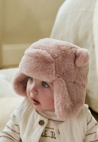 Rožinės spalvos dirbtinio kailio medžioklinė kepurė su ausų lopinėliais, minkšta tekstūra, apvaliu viršumi ir mažomis meškiuko ausytėmis. Turi prigludimą ir šiltą dizainą.