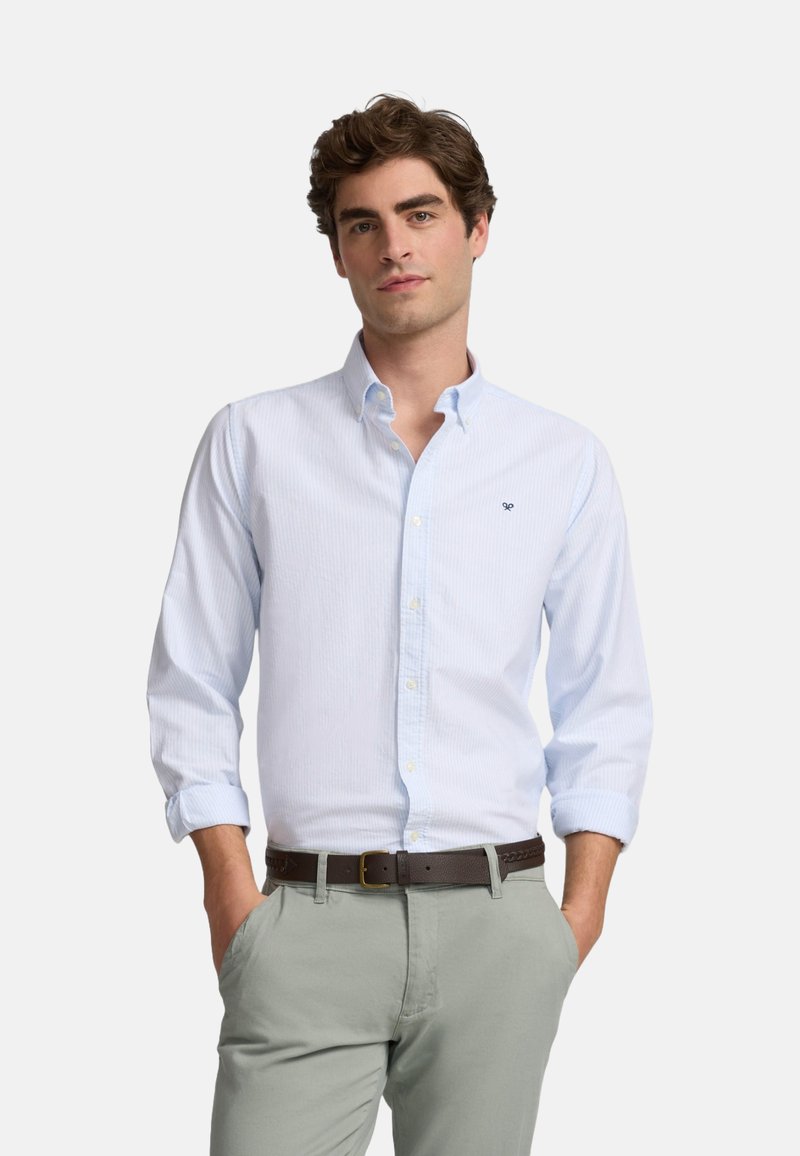 Camisa a rayas azul claro, con cuello, cierre de botones y mangas remangadas. Lleva puesta un pantalón gris claro y un cinturón marrón.