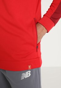 Rød atletisk langærmet pullover i struktureret stof med kontrasterende maroon ærmeaccenter, sidelomme med lynlås og broderet LFC-logo.