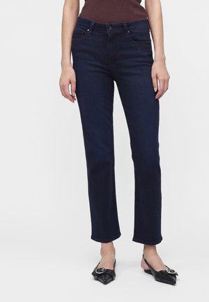 Frau trägt dunkelblaue Jeans mit geradem Bein und schwarze Slingback-Ballerinas mit spitzer Spitze und Schnallendetail vor einfachem Hintergrund.