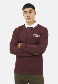 Polo a maniche lunghe color bordeaux con colletto bianco, con la scritta "COPENHAGEN EST. 1972" ricamata sul petto. Tessuto di cotone morbido.
