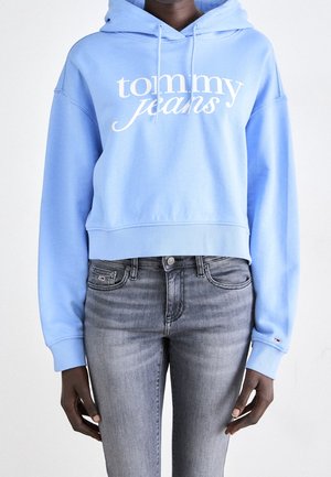 Personne portant un sweat à capuche bleu clair Tommy Jeans et un jean gris ajusté, debout les bras détendus le long du corps sur un fond uni.