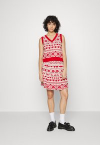 Vestido sem mangas em vermelho e bege com padrões, apresentando um decote em V, com motivos geométricos e corações, terminado com um cinto sólido vermelho.