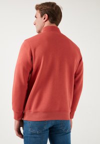 Sudadera roja con un cuello alto y puños acanalados, hecha de una tela suave. Tiene un corte relajado y presenta una textura lisa.