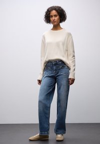Maglione crema con polsini a coste, abbinato a jeans blu a vita alta e gamba larga con tasche frontali. Scarpe color cammello completano l'outfit.