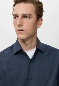 Camisa de botones de color azul marino oscuro con un patrón texturizado y cuello extendido, usada sobre una camiseta blanca de cuello redondo, con botones negros.