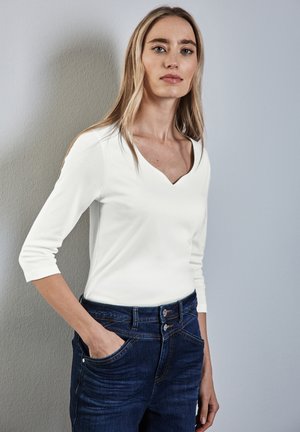 Maglietta bianca a maniche lunghe con scollatura a V, abbinata a jeans blu scuro. Presenta una texture liscia e una silhouette aderente.
