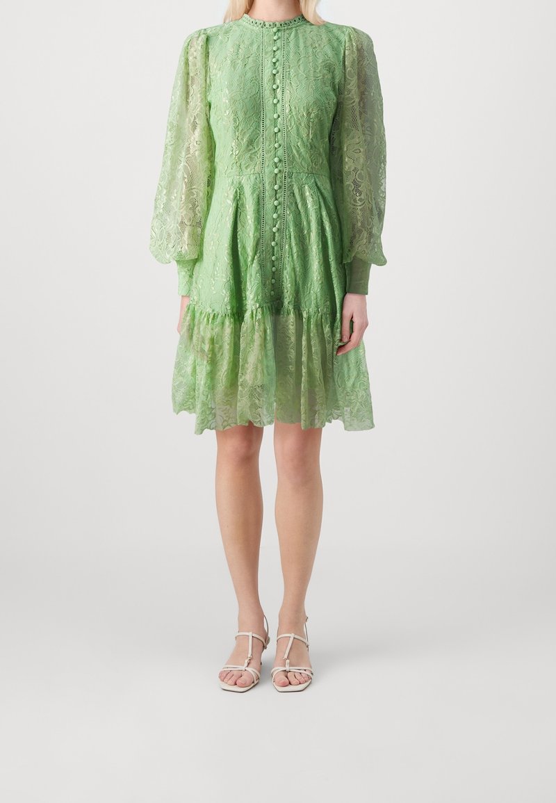 Femme portant une robe en dentelle vert clair avec de longues manches bouffantes et des boutons sur le devant, associée à des sandales blanches à brides.