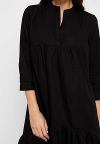 Musta puuvillamekko korkealla kauluksella, kolme neljäsosaa hihat ja kootut kerrokselliset helmat. Sileä rakenne, minimalistinen muotoilu.