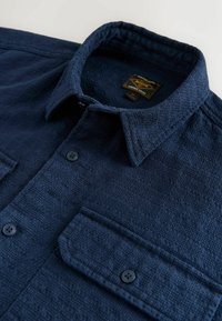 Chemise texturée bleu marine avec un col boutonné, deux poches poitrine et des boutons noirs. Le tissu présente un motif tissé pour plus de profondeur.