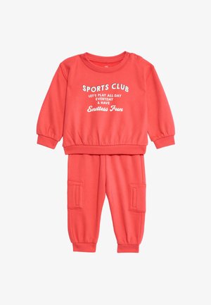Korálový sportovní outfit s dlouhými rukávy a kalhotami. Obsahuje žebrované manžety, elastický pas, přední kapsy a bíle tištěný text.