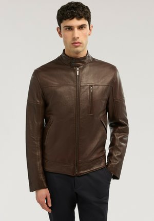 Joven con chaqueta de cuero marrón oscuro con cremallera, cuello tipo mao y bolsillo en el pecho con cremallera, posando frente a un fondo liso y claro.