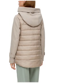 Femme portant une veste beige avec un dos matelassé et des manches en tissu, associée à un pantalon vert clair, vue de dos sur fond blanc.