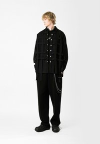 Camisa negra de gran tamaño con detalles de tirantes y botones, combinada con pantalones holgados negros y zapatos negros texturizados. Fondo liso.