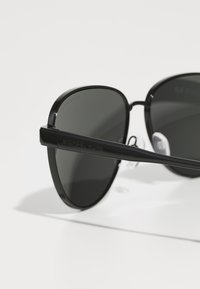 Lunettes de soleil noires avec une monture lisse et arrondie, et des verres foncés. Présente un logo subtil sur la branche et des plaquettes de nez transparentes.