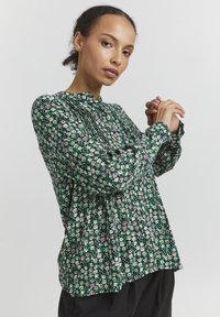 Blusa floreale verde caratterizzata da piccoli fiori bianchi, rosa e verdi, con colletto alto e design plissettato, maniche lunghe e vestibilità ampia.