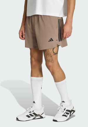 Mężczyzna ubrany w brązowe sportowe szorty, białe skarpetki Adidas oraz białe buty sportowe Adidas, z widoczną tatuażowaną prawą nogą.