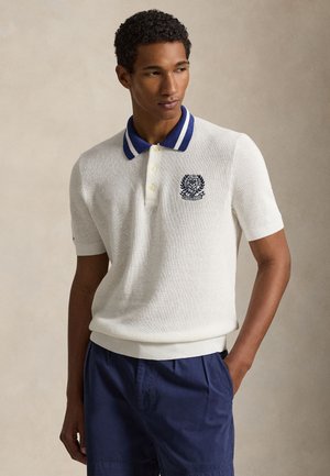 Ung mann iført en hvit strikket polo med blå krage og Wimbledon-logo, kombinert med marineblå shorts, stående med den ene hånden i lomma.