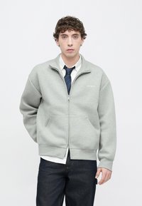 Felpa grigia con cerniera, tasche frontali, polsini e collo a coste; indossata sopra una maglia bianca a maniche lunghe e jeans scuri.