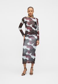 Γυναίκα με μακρυμάνικο, εφαρμοστό φόρεμα με λουλούδια σε αποχρώσεις μαύρου, λευκού και μπουρντό, φορώντας μαύρα ψηλοτάκουνα σανδάλια και ασημένια τσάντα ώμου.