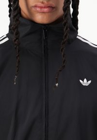 Črna jakna z zadrgo, izdelana iz lahkega materiala. Ima bel tri-stripe vzorček na ramah in majhen bel logotip na prsih.