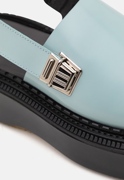 Lyseblå syntetisk rem sandal med sort platform sål, med en metallisk spænde detalje og strukturerede indslag langs sålen.