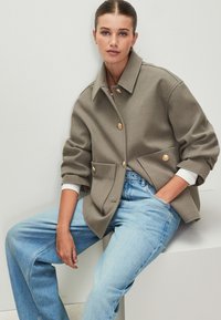 Olivzaļš oversize mētelis ar apkakli un priekšējām kabatām, rotāts ar zelta pogām. Gaiši zilas, augsta vidukļa džinsi ar brīva piegriezuma stilam.