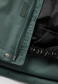 Grøn vejrbestandig jakke med lynlås, der har en glat tekstur, broderet logo og en sort inderside med et mærket bånd.