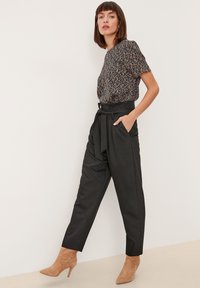 Donna in piedi contro un muro bianco, indossa una blusa a maniche corte con fantasia, pantaloni neri a vita alta con cintura a fiocco e stivali con tacco color marrone chiaro.