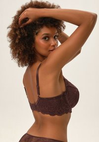 Brązowy koronkowy biustonosz z kwiecistym wzorem, regulowanymi ramiączkami i zapięciem na hak z tyłu. Modelka pozuje bokiem z kręconymi włosami.