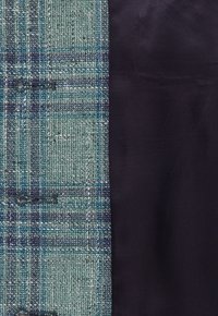 Tweedtyg till vänster har blå och gröna rutig mönster. Höger sida är i ett slätt, mörknavy material. Synliga knappar som är markerade längs sömmen.