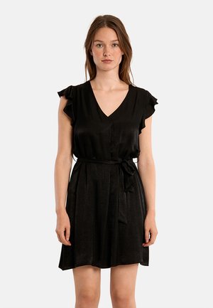 Femme debout portant une courte robe noire avec des manches courtes à volants et une ceinture nouée à la taille sur un fond blanc uni.