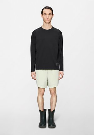 Mann steht vor weißem Hintergrund, trägt ein schwarzes Langarmshirt, helle Shorts und schwarze Gummistiefel, schaut nach vorne mit neutralem Gesichtsausdruck.