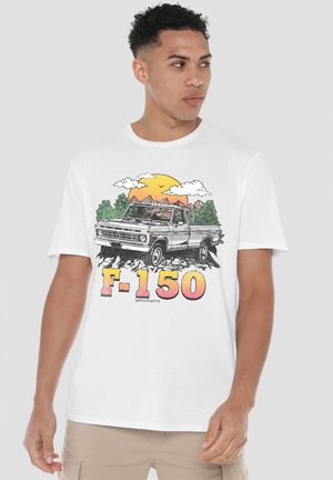 Man wearing white T-shirt with vintage F-150 truck illustration, sunset, trees, and mountains, looking sideways.
