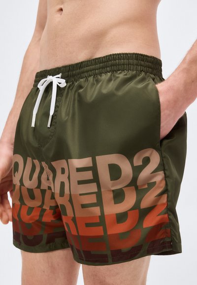 Άνδρας που φοράει μαγιό σε ελιά πράσινο με μεγάλο κείμενο «DSQUARED2» σε μπεζ και κόκκινο, με ελαστική μέση και λευκό κορδόνι, το χέρι στην τσέπη.