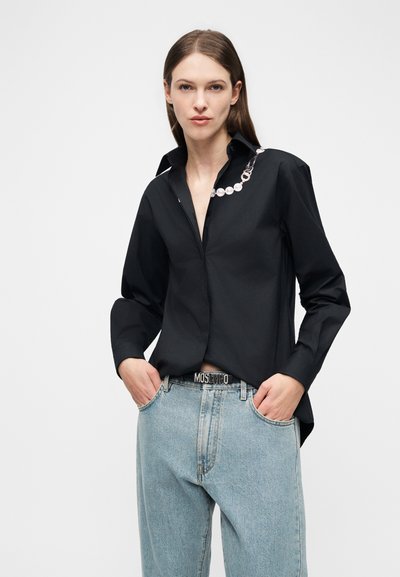 Γυναίκα που φοράει μαύρο μπλουζάκι με μακριά μανίκια και λεπτομέρεια αλυσίδας με πέρλες, καθώς και ανοιχτό γαλάζιο ψηλόμεσο τζιν με ορατή ζώνη Moschino.