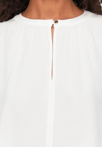Witte katoenen blouse met een ronde halslijn, een knoopsluiting aan de voorkant en subtiele plooien langs de halslijn.