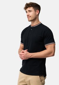 Crna polo majica s ovratnikom na gumbe, koja sadrži kontrastne prugaste detalje na ovratniku i rukavima. Glatka tekstura tkanine.